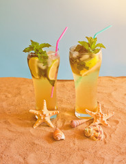 Two glasses of mojito cocktail with drinking straws on sand beach on blue backdrop.