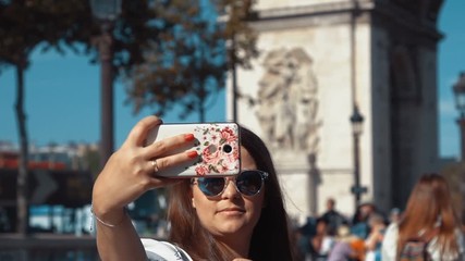 Professional video of beautiful female taking selfie while on urban street with Arc de Triomphe behind her. Paris in slow motion