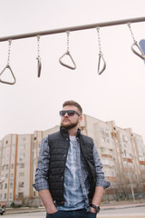 A man with a beard and sunglasses in a plaid shirt and waistcoat posing on the street to advertise...
