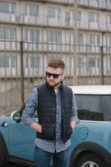 A man with a beard and sunglasses in a plaid shirt and waistcoat posing on the street to advertise...