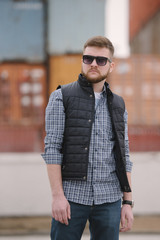 A man with a beard and sunglasses in a plaid shirt and waistcoat posing on the street to advertise...