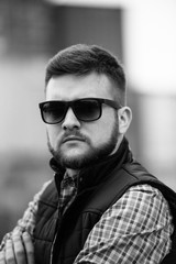 A man with a beard and sunglasses in a plaid shirt and waistcoat posing on the street to advertise men's clothing. Advertising menswear