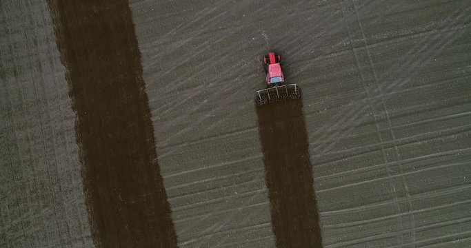 Red tractor with harrows during plowing brown field with green grass around. Top down drone view in uhd.