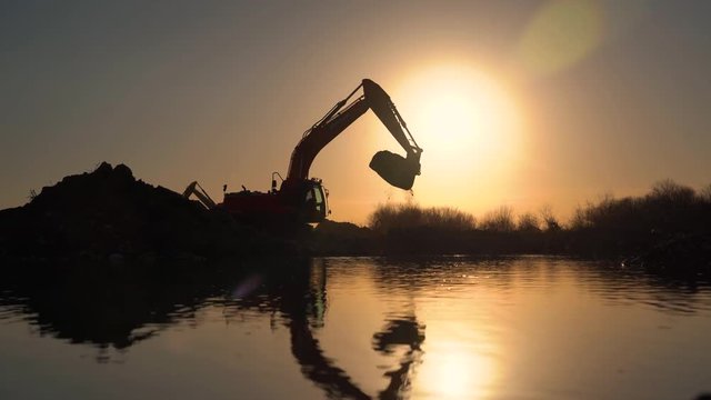 Silhouette of excavator. Dredge digs ground and loading tipper. Digger machine digging hole. Excavation of canal. Dump truck,tracked,bagger,backhoe,shovel,sunny blue sky,sun in background,backlight.
