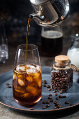 Ice coffee cold summer drink in a tall glass with pouring coffee and coffee beans on a stone background.