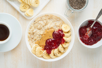 Top view of oatmeal porridge with strawberry jam, peanut butter, banana, coffee on white wooden light background, healthy vegan diet breakfast