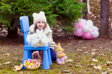 Outdoor photo of cute little girl with real Easter bunny sitting on the blue chair in front of her.Easter concept