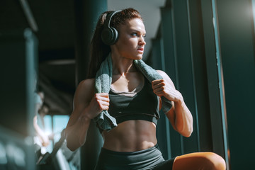 Young attractive strong muscular female bodybuilder with ponytail and headphones posing in gym with towel around neck. Dreams don't work unless you do.