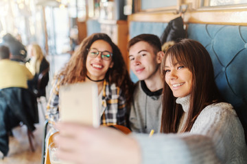 Three multicultural teenage classmates taking selfie while sitting at cafeteria.