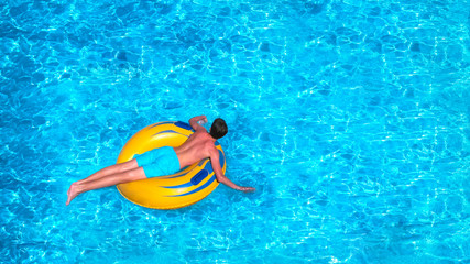 Young man is floating on yellow inflatable air ring/circle in pool with blue water. Holiday leisure in happy sunny day. Vacation concept, top view.