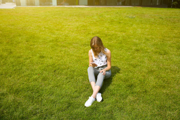 Beautiful hipster girl is sitting near modern architecture building. Stylish business woman looking in the smartphone on green grass lawn. Student surfing in networks. Lifestyle relax. Office worker.