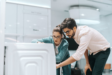 Attractive multicultural couple looking at new washing machine they want to buy. Tech tore interior.