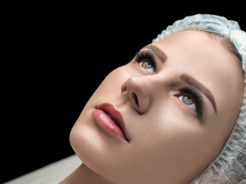 Eyelash extension result. Female face with long black lashes and disposable cap on couch in beauty saloon, selected focus.