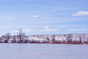 Lake bank covered with ice, trees without leaves on hill, blue cloudy sky background
