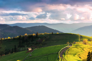 mountainous countryside in evening light. grassy rural fields on rolling hills. wooden fence along the path. mountain ridge in the distance. wonderful weather with pink fluffy clouds