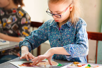 Fototapeta premium Portrait of blonde girl with big black glasses smiling happily while enjoying art and craft lesson in art school working together with other kids