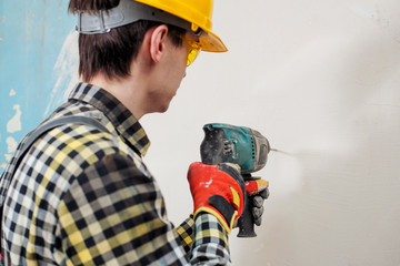 A young builder in a yellow helmet with a perforator, drills the wall in the room for repair.
