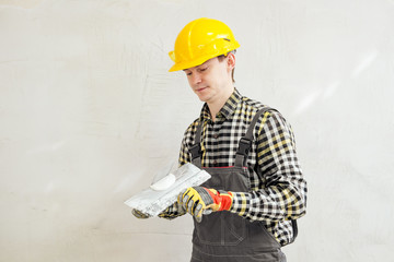 Young male builder and repairman in a yellow helmet plastering the wall with a spatula and plaster.