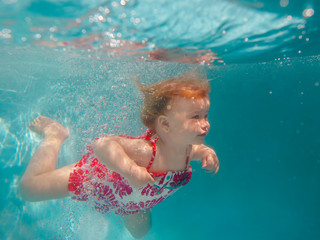 Obraz na płótnie Canvas Smiling baby girl in cute modern dress diving underwater in blue swimming pool. Active lifestyle, child swimming lesson with parents. Water sports activity during family summer vacation