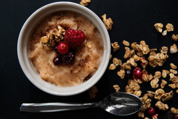 Photo of porridge with berries on top, served in a ceramic pie dish and placed onto a black table/ background, with oatmeal and berries over the place.