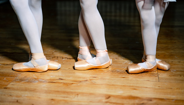 Feet of a three young ballerinas in pointe shoes against the background of the wooden floor. Legs in white tights and beige pointes in dancing studio. Close-up