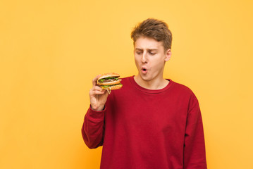 Funny young man holds a burger in his hands, looks at fast food with a surprised look, isolated on a yellow background. Hungry teenager with burger in his hands is on a yellow background.