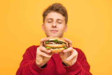 Handsome guy with his eyes closed shows a delicious large burger to the camera, holds in his hands, isolated on a yellow background.Focus on an appetizing burger in the hands of a young man. Fast food
