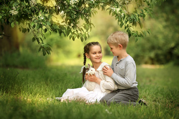 Children sitting under a blooming apple tree in a summer park play with a small cute rabbit