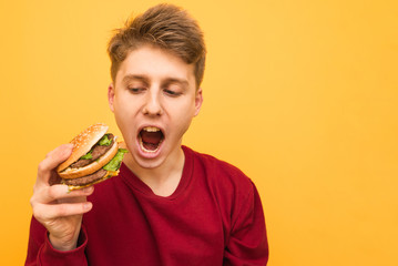 Portrait of an emotional young man with a burger in his hands on a yellow background. A hungry emotional guy holds a burger in his hands and shouts, isolated. Very hungry student. Copyspace