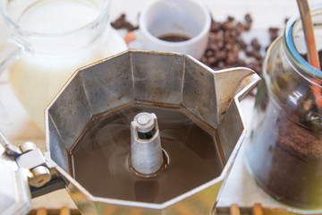 Teapot with some black coffee on a wooden table next to coffee beans, milk and a cup in a rustic kitchen.
