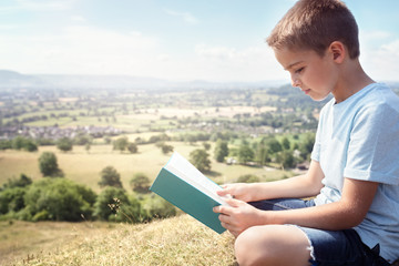 Boy sitting on a hill reading a book in a meadow