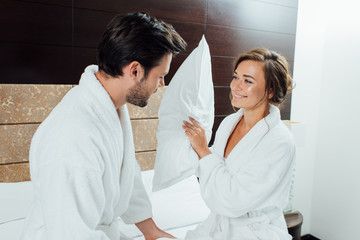 Fototapeta premium happy couple in white bathrobes having pillow fight on bed in hotel