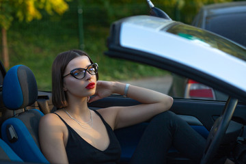 Pretty brunette model wearing glasses posing in a convertible car with natural soft light. Space...