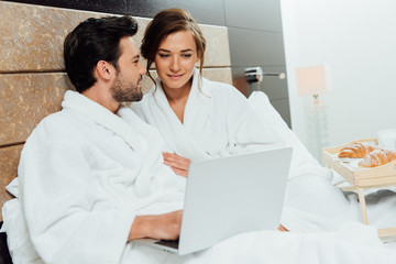 beautiful woman holding cup and looking at laptop near handsome bearded man in bed