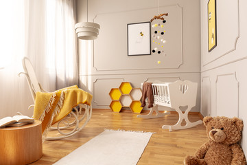 Yellow blanket on white wooden rocking chair in spacious baby bedroom with teddy bear and rug on...