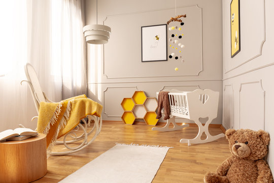 Yellow blanket on white wooden rocking chair in spacious baby bedroom with teddy bear and rug on the floor