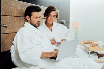 Obraz premium beautiful woman holding cup and looking at laptop with handsome man in bed
