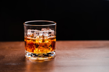 Glass of whiskey with ice cube on wooden table over black background