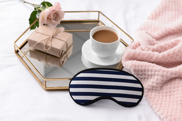 Tray with cup of coffee, flower, gift and sleep mask on bed