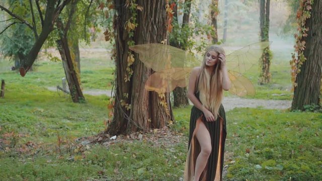attractive fairy with blond hair dancing to rhythm of spring forest singing and gently straightening her blond hair, nymph in green dress with cuts in hem for bare legs, smiling tenderly at camera