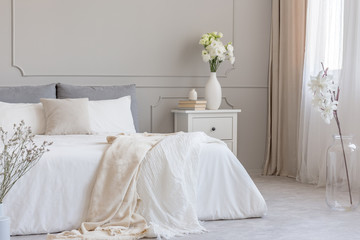 Roses in white stylish vase and pile of books on wooden nightstand with drawers next to cozy bed with blanket
