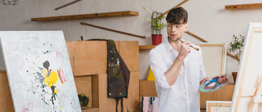 panoramic shot of handsome artist with paintbrush and palette looking at painting in painting studio