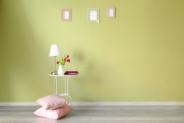 Table with tulips, lamp and pillows near color wall in room