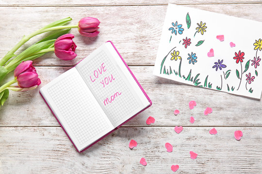 Beautiful picture with tulips and notebook with text LOVE YOU MOM on wooden table