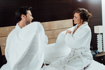cheerful couple in bathrobes having pillow fight on bed in hotel