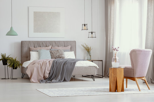Flowers in glass vase on wooden coffee table next to stylish pastel pink armchair in elegant bedroom design with double bed with headboard and cozy bedding