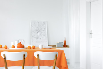 Table with oranges, pumpkin, dinnerware and juice placed in white dining room interior with cupboard with books and map poster in the real photo