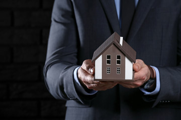 Insurance agent with model of house on dark background, closeup. Protection of house from earthquake