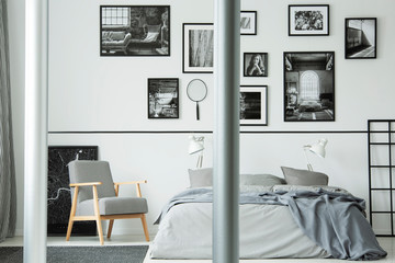 Wooden armchair next to bed in white bedroom interior with gallery of photos on the wall. Real photo