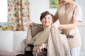 Young nurse in beige uniform helping smiling senior patient on wheelchair in nursing home